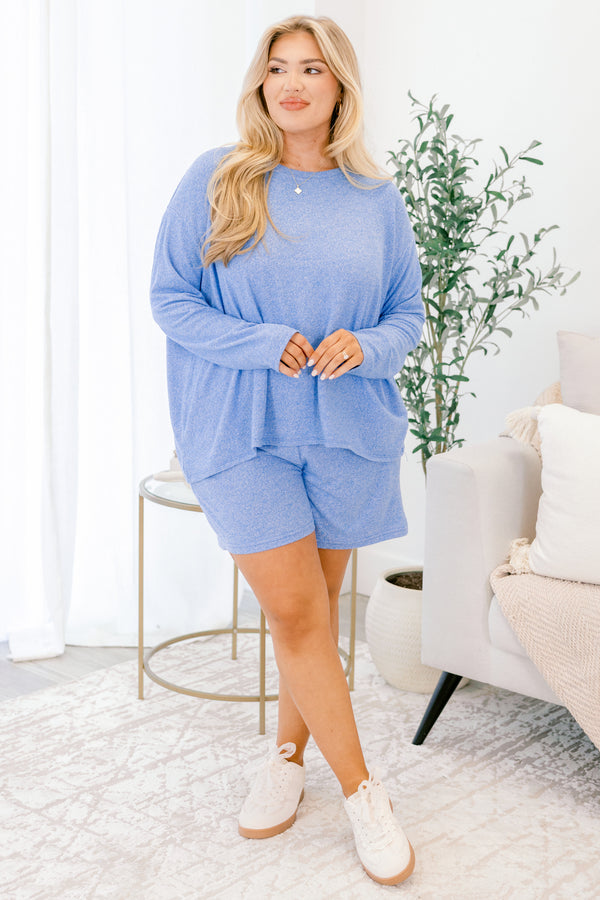 Woman wearing a blue outfit standing in a bright room with a plant and chair.