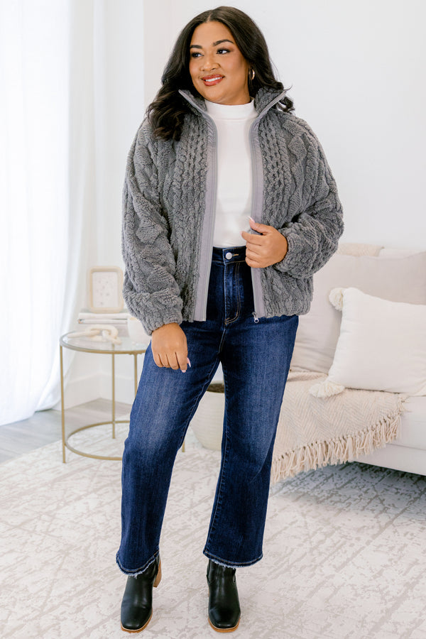 Woman wearing a gray cable knit sweater, white shirt, blue jeans, and black boots in a living room.