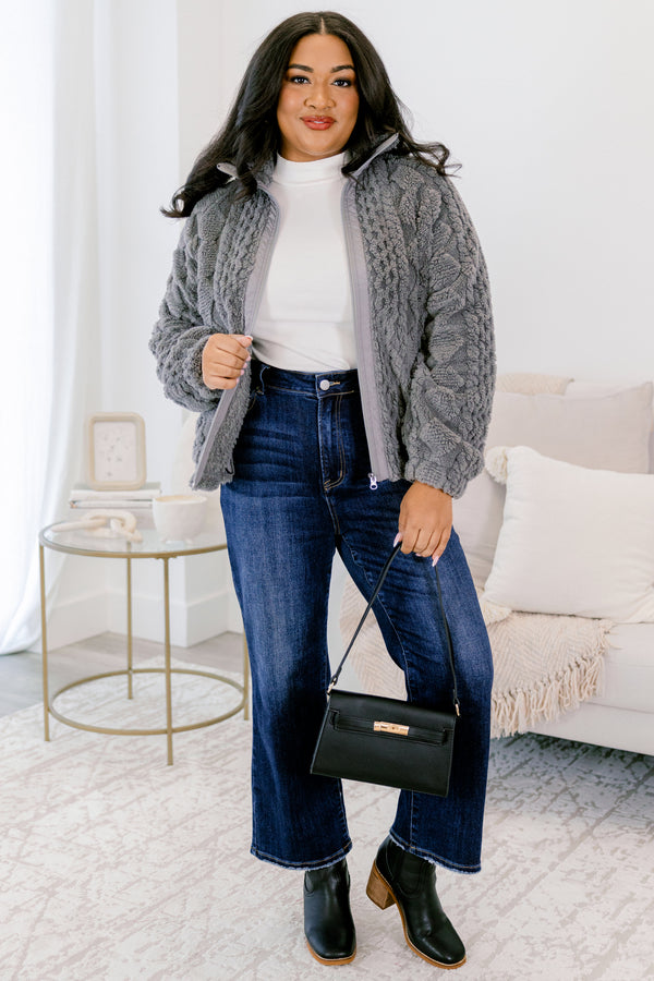 Woman wearing a gray sweater, white shirt, blue jeans, and black boots in a living room.