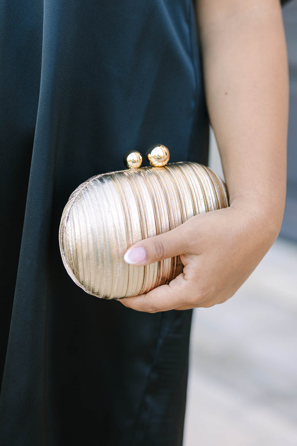 A woman holding a gold textured oval clutch with a gold chain strap and ball clasp.