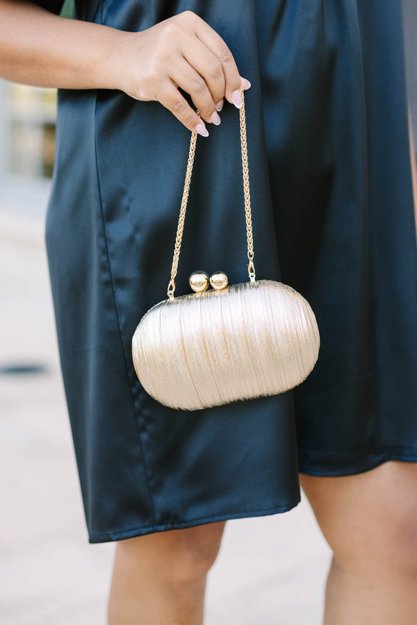 A woman holding a gold textured oval clutch with a gold chain strap and ball clasp.