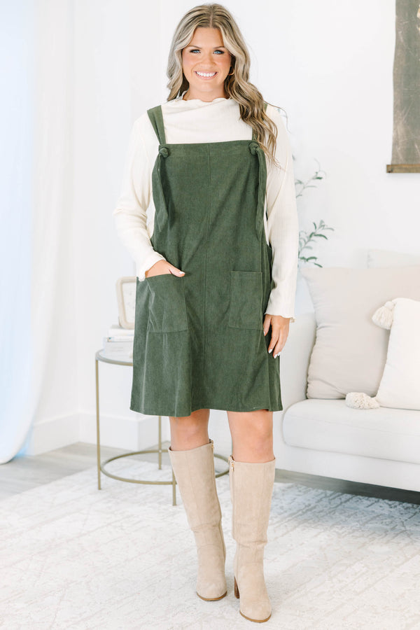 Plus size olive green corduroy overall dress with tie straps and front pockets over a cream top.