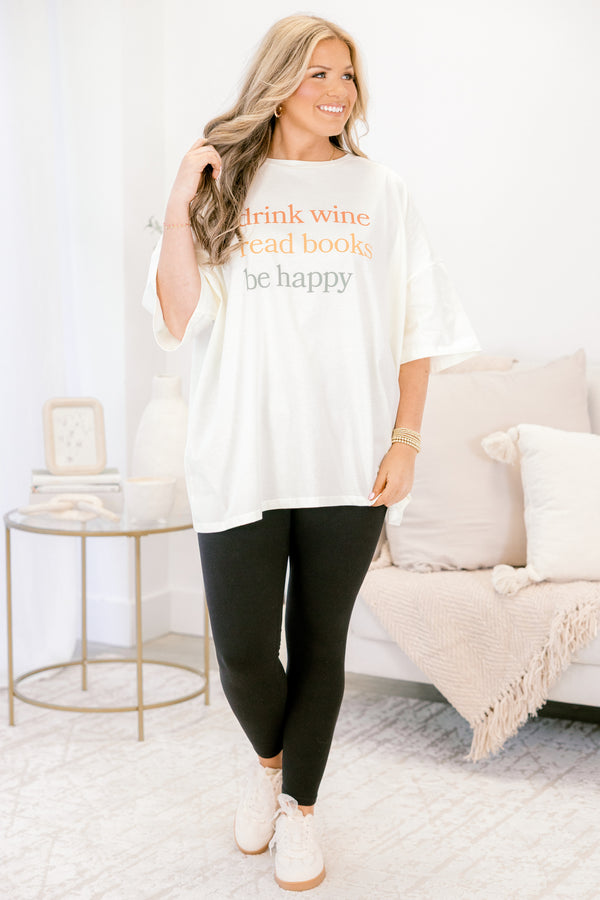 Woman wearing a white oversized t-shirt with text, black leggings, and white sneakers in a living room.