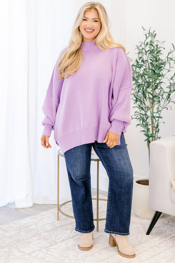 Woman wearing a purple sweater and blue jeans standing in a room with a plant and chair.