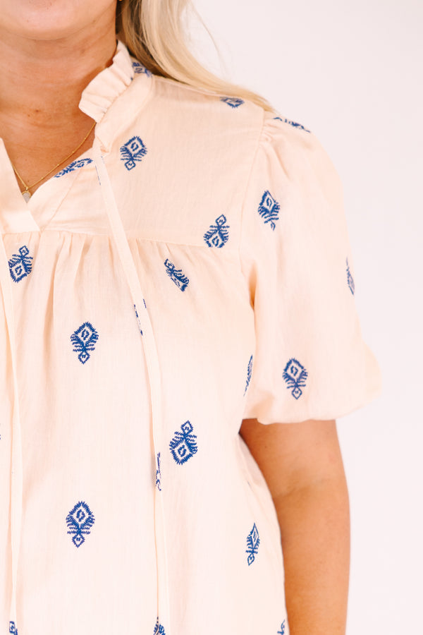 Chic Soul plus size clothing, Beige short-sleeve blouse with a relaxed, flowy fit, featuring blue embroidered geometric patterns, a ruffled split neckline with tie tassels, and a decorative blue border along the hem.