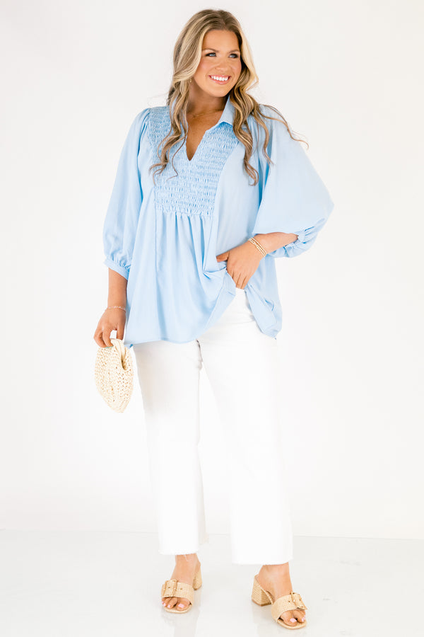 Light blue blouse featuring a split neckline, smocked front detail, and long sleeves with soft gathered cuffs in a lightweight, flowy fabric.