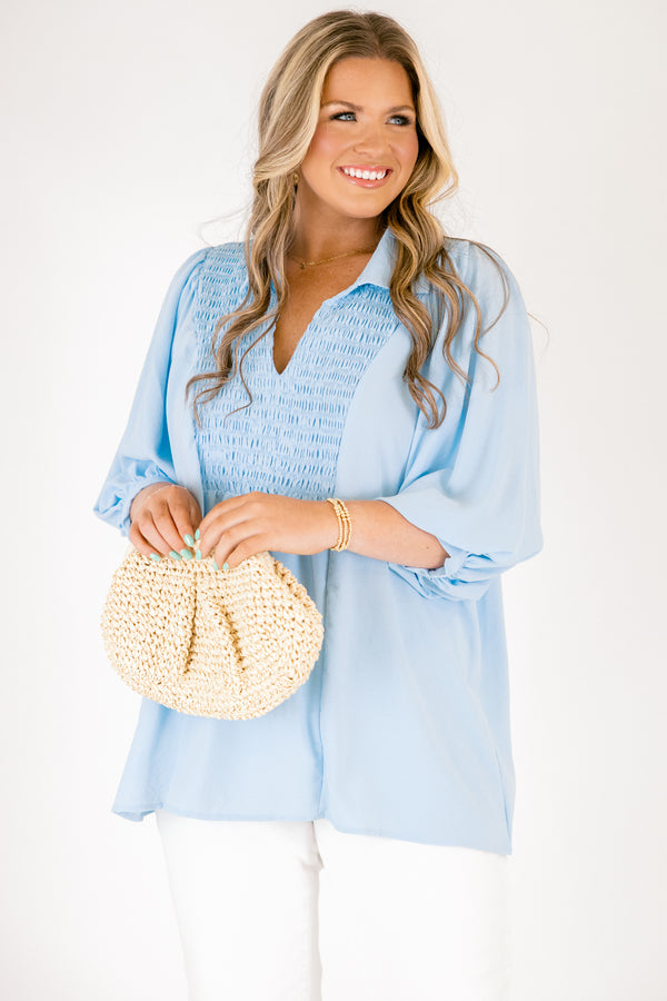 Light blue blouse featuring a split neckline, smocked front detail, and long sleeves with soft gathered cuffs in a lightweight, flowy fabric.